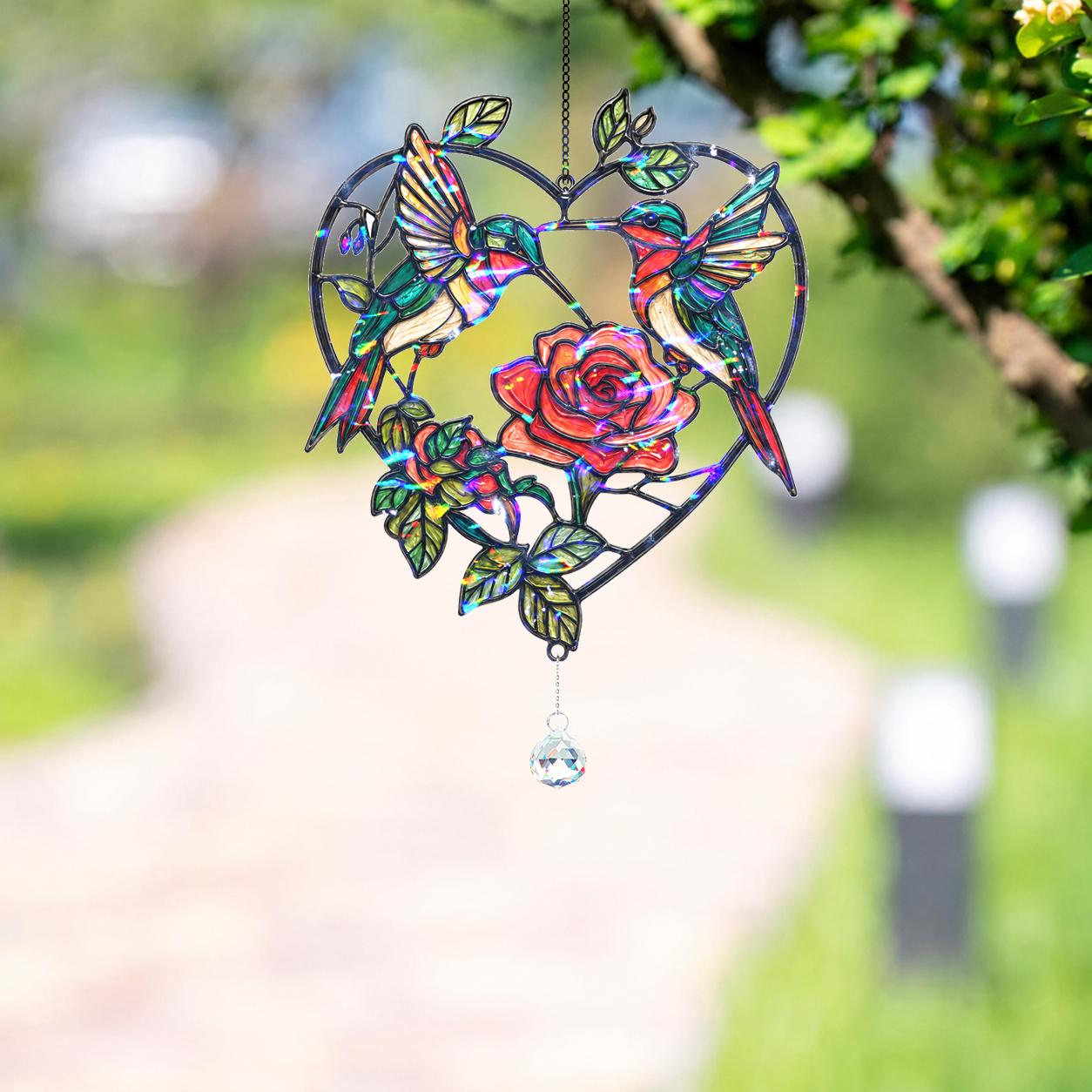 Two hummingbirds rose heart holographic acrylic suncatcher hanging outdoors with a soft green garden background.
