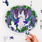 Hand placing a puzzle piece into the Wisteria Duet Hummingbird acrylic puzzle suncatcher with loose pieces nearby.